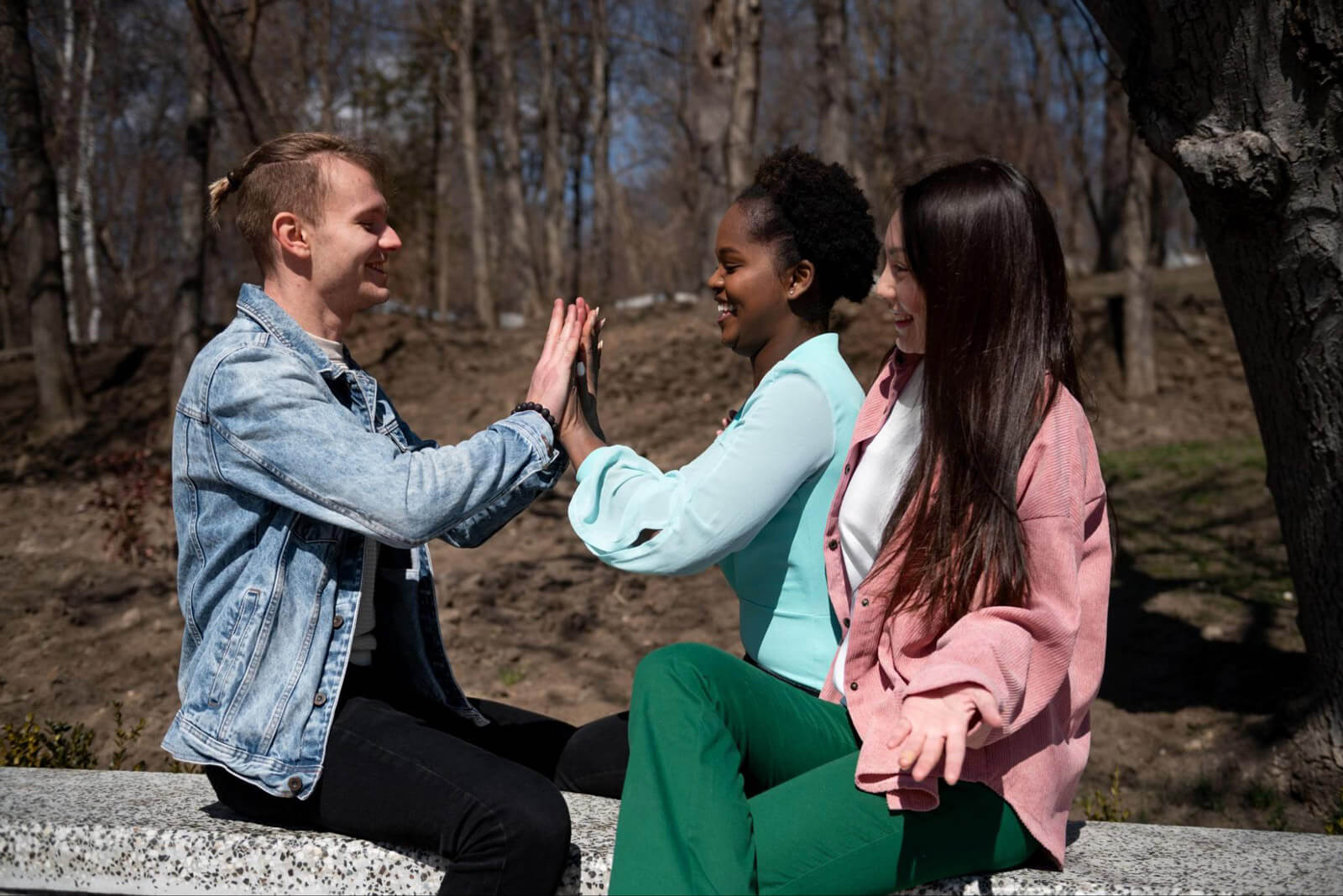 Three young adults sit together on a stone bench in a wooded park, smiling as two of them share a supportive high-five.