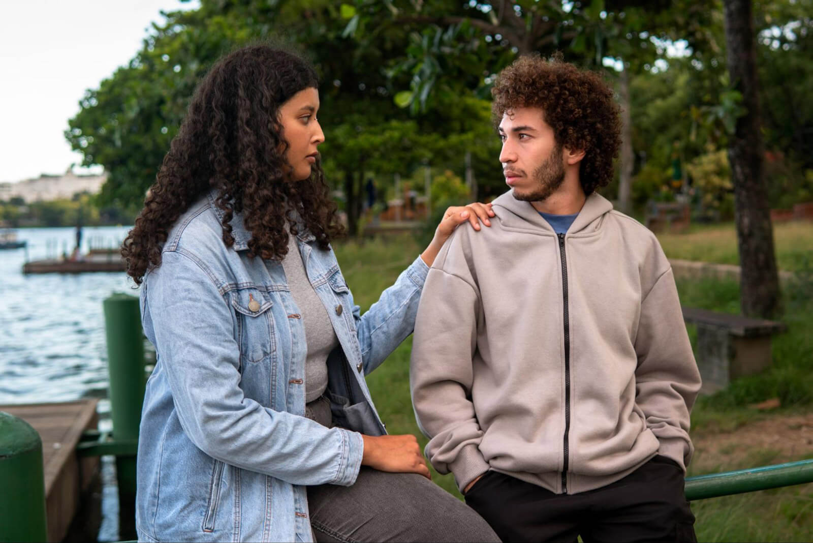 Two young adults stand beside a lakeside path while one places a reassuring hand on the other’s shoulder during a thoughtful conversation.